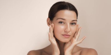 Yüzüne nazikçe dokunan parıldayan, doğal ciltli genç bir kadının yakın çekimi. Yumuşak ışıklandırma onun sağlıklı tenini güçlendirir, bu görüntüyü güzellik, cilt bakımı ve sağlık reklamları için ideal kılar.