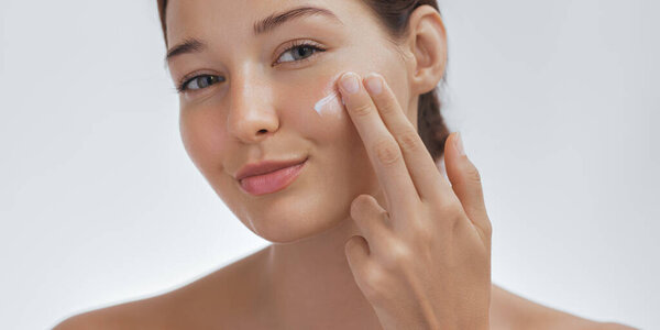 Женщина с сияющей, светящейся кожей нежно наносит крем для кожи на лицо. Мягкое освещение и безупречный цвет лица делают этот образ идеальным для красоты, кожи, и оздоровительные акции сосредоточены