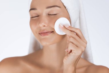 Havlusu olan sakin bir kadın yüzünü pamukla siliyor, pürüzsüz ve pürüzsüz bir cildi var. Güzellik, cilt bakımı ve sağlıklı, parlayan cilde odaklanmış sağIık için mükemmel..