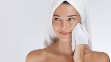 A happy woman with a towel wrapped around her head cleanses her face with a wipe, enjoying her skincare routine. Perfect for illustrating concepts of beauty, self-care, hygiene, and fresh skin care. clipart