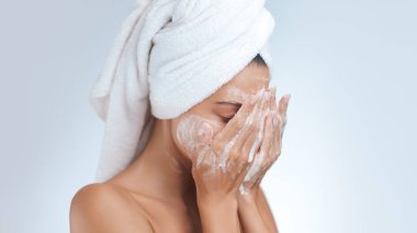 A woman with a towel wrapped around her head gently cleanses her face with foaming soap during her skincare routine. Ideal for concepts related to beauty, self-care, hygiene, and healthy skin clipart