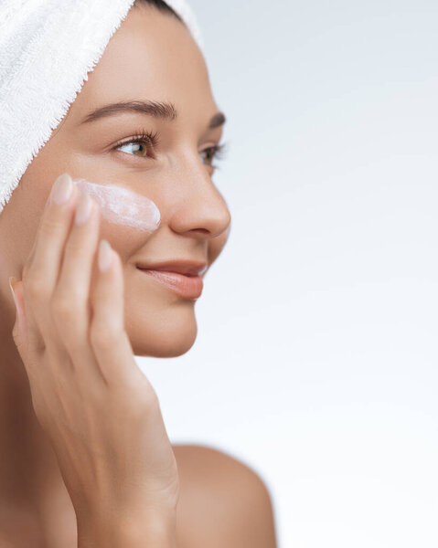 Крупный план улыбающейся женщины с полотенцем, обернутым вокруг головы, наносящей крем для кожи на щеку, наслаждаясь преимуществами своей рутины по уходу за кожей. Перфект для иллюстрации тем красоты, себя