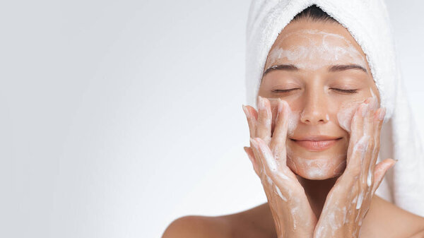 Portrait of a young woman with towel-wrapped hair gently applying foam cleanser to her face. Ideal for promoting skincare, wellness products, beauty treatments, or spa services in an elegant