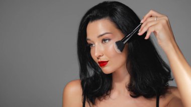 Makyaj fırçası kullanarak çarpıcı kırmızı dudakları olan, kusursuz cilt ve güzellik rutinlerini vurgulayan göz kamaştırıcı bir kadının yakın çekimi. Kozmetik, moda yazıları ve cilt tanıtımları için ideal