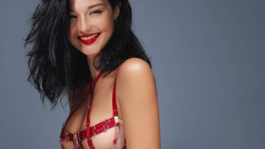 Kırmızı iç çamaşırlı kendine güvenen bir kadın parlak bir şekilde gülümsüyor, koyu kırmızı rujuyla vurgulanıyor. Koyu renk saçları canlı iç çamaşırlarıyla çelişiyor. Bu da bu görüntüyü moda, güzellik ve iç çamaşırı için ideal kılıyor.