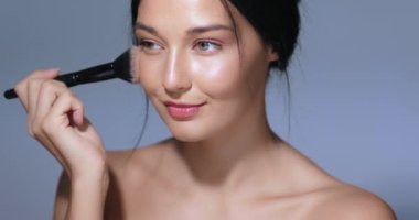 Parlak ciltli ve makyajlı güzel bir kadın makyaj fırçasıyla allık sürüyor, stüdyo ortamında gülümsüyor. Güzellik dersleri, cilt bakımı reklamları ve kozmetik reklamları için mükemmel.. 