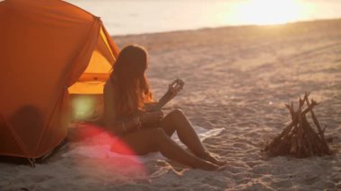 Sahilde mükemmel bir yaz anının tadını çıkaran bir boho kızı gün batımında çadırının yanında ukulele çalıyor. Rahatlama, seyahat ve tasasız yaşam tarzları için idealdir..