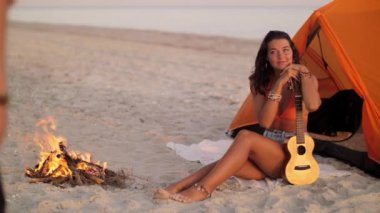 Çadırlarının yanında kamp ateşi yakan bir çiftin olduğu romantik bir plaj kampı akşamı. Kadın ortağına gülümserken bir gitar tutuyor. Seyahat, eğlence ve açık hava macerası için mükemmel.