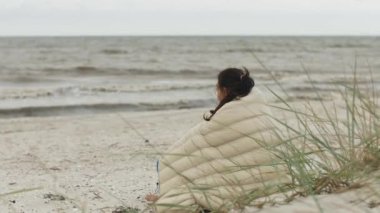 Kumlu bir sahilde battaniyeye sarılmış bir kadın bulutlu bir günde okyanusa bakıyor, sakin ve düşünceli bir anın tadını çıkarıyor. Sıcak bir battaniyeye sarılmış bir kadın kumlu bir sahilde oturur ve bakar.