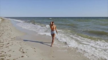 Güneşli bir sahilde, bikinili bir bluz ve kot şort giymiş, sıcak havanın ve okyanus dalgalarının tadını çıkaran bir kadın. Bikini bluzlu ve şortlu bir kadın güneşli plajda geziniyor.