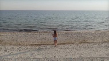 Kumlu bir plajın kıyı şeridi boyunca denize koşan bir kadının sinematik drone görüntüleri. Seyahat blogları, yaz promosyonları ve yaşam tarzı içeriği için mükemmel. 