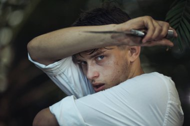 Yoğun bir ormanda, kirli ve yorgun bir şekilde bıçaklanmış, gergin bir genç adam. Tehlike, hayatta kalma ve dayanıklılık temalarını yakalar. Yüksek kaliteli promosyon malzemeleri için idealdir.