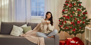 Bir kadın, yanan bir Noel ağacının yanındaki koltukta rahat bir şekilde otururken sıcak bir içkinin tadını çıkarır. Bu resim, tatil temalı pazarlama veya gevşeme içeriği için ideal samimi ve neşeli ruhu yakalar.