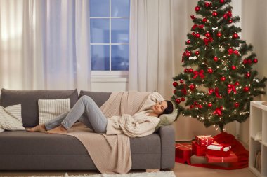 Huzurlu bir kadın, gözleri yanan bir Noel ağacının yanında kapalı bir kanepede uzanıp sıcak ve şenlikli bir ortam yaratıyor. Tatil temalı reklamlar ya da rahatlama odaklı promosyonlar için mükemmel..