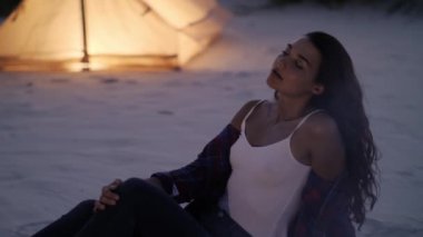 Huzurlu genç bir kadın, arka planda sıcak bir çadırı olan kumlu bir kamp alanında, parlayan bir kamp ateşinin yanında oturur. Açık hava rahatlığının ve huzurlu kaçışların özünü yakalar..