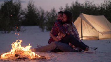 Romantik çift, geceleri kumlu bir kamp alanında çadırlarının yanında parlayan bir kamp ateşinin yanında sarılıyorlar. Aşk, açık hava macerası, kamp ve huzurlu kaçamaklar için mükemmel bir tema.. 