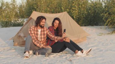 Çadırlarının dışında kumlu bir sahilde oturan mutlu çift akıllı telefon kullanarak bir video görüşmesi yapıyor. Rahatlatılmış dış mekan bağlantıyı, macerayı ve doğayı ön plana çıkarıyor. Seyahat ve yaşam tarzı için ideal.