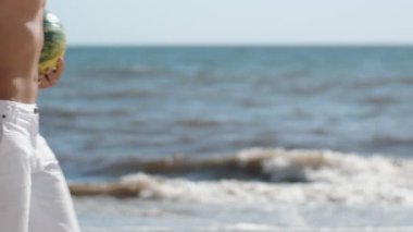 Kumsalda yürüyen bir çiftin yakın plan çekimi. Arka planda okyanus olan örülmüş bir plaj çantası taşıyan bir kadın ve elinde karpuz tutan bir adam. Yaz temaları için mükemmel