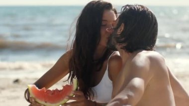 Romantik bir çift, arka planda okyanus olan karpuz dilimlerini tutarken güneşli bir sahilde tatlı bir öpücük paylaşır. Aşk, yaz eğlencesi, sağlıklı yaşam ve romantik kavramları için mükemmel.