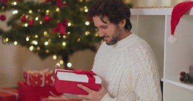 Noel hediyesini Noel ağacıyla süslenmiş bir salonda açan, rahat örgü süveterli gülümseyen bir adam. Tatil promosyonları, şenlikli reklamlar ve mevsimsel içerik için mükemmel..