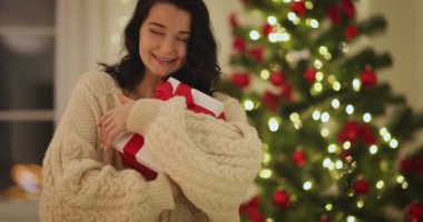 Güzel dekore edilmiş ışıklarla süslenmiş bir ağacın yanında kırmızı ve beyaz bir Noel hediyesini tutarken gülümseyen rahat bir süveter giymiş bir kadın. Tatil promosyonları ve festival reklamları için mükemmel..