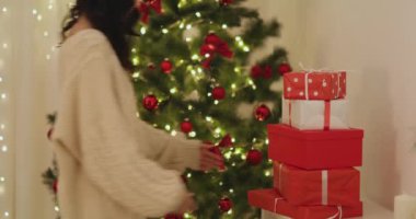 Hoş bir süveter ve Noel Baba şapkası giymiş neşeli bir kadın güzel süslenmiş bir ağacın önünde bir yığın Noel hediyesiyle sıvışıyor. Bayram temalı promosyonlar ve şenlikli pazarlama için mükemmel..