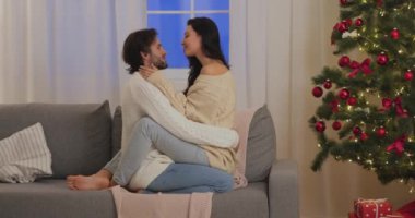 Rahat kazaklı genç çift romantik bir anı paylaşıyor ve bir Noel ağacıyla şenlikli bir oturma odasında kanepede kucaklaşıyorlar. Tatil promosyonları, aşk temalı içerik ve...