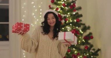 Noel Baba şapkası takan neşeli bir kadın ve elinde Noel hediyeleri tutarken rahat bir kazak dans ediyor. Sahne güzel aydınlatılmış ve dekore edilmiş bir Noel ağacının önünde geçiyor. Festival için ideal.