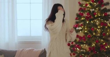 Neşeli bir Asyalı kadın, güzel dekore edilmiş bir Noel ağacının yanında rahat bir kazakla dans ediyor, neşeli ruhu somutlaştırıyor. Tatil, kutlama ve yaşam tarzı temaları için mükemmel. Yüksek kaliteli görüntüler