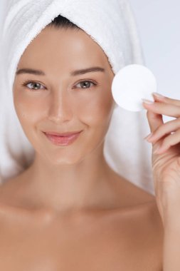 Kafasına havlu sarılmış bir kadın elinde pamuk tutarak gülümsüyor ve parlayan tenini parlaklaştırıyor. Taze ve sağlıklı cilt bakımını vurgulayan cilt bakımı, güzellik ve sağlık kampanyaları için ideal