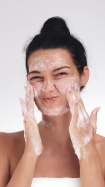 Neşeli genç bir kadın yüzünü köpükle yıkıyor, sevimli bir ifade takınıyor. Cilt bakımı, güzellik ürünleri ya da temiz, parıltılı ve ışıl ışıl cilt bakım rutinlerine odaklanan sağlık reklamları için idealdir..