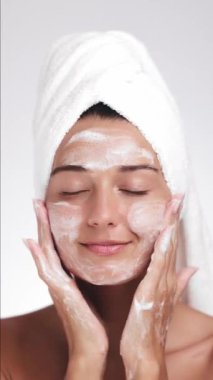 Havluyla sarıp sarmalayan ışıl ışıl bir kadın yüzünü köpüklü temizleyiciyle nazikçe temizliyor, gözleri kapalı gülümsüyor. Cilt bakımı, güzellik ya da sağlıklı ürün promosyonları için mükemmel.. 
