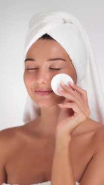 Pamuk ped kullanan, parlayan derisi ile gülümseyen, havluyla sarıklı parlak bir kadın. Tazelik ve zarafete odaklanan güzellik, özbakım veya cilt ürünlerini teşvik etmek için idealdir. 