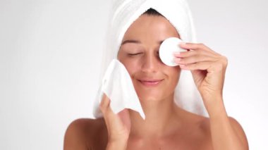 Kafasına havluyla sarılmış gülümseyen bir kadın cilt bakımı sırasında pamuk pedi ve yüz bezi kullanıyor. Güzellik, özgüven ve neşe kavramlarını resmetmek için mükemmel.