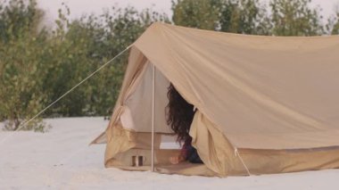 Genç bir kadın kumlu bir sahilde çadırından dışarı bakıyor ve güne bir kamp gezisi sırasında başlıyor. Bu görüntü doğada uyanmanın heyecanını yakalar. Macera ve seyahat için mükemmeldir.