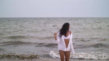 Beyaz bikinili ve açık tişörtlü bir kadın gri gökyüzünün altında fırtınalı bir okyanusun kenarında duruyor. Dramatik hava ve karamsar atmosfer yalnızlık ve doğal güzellik temaları yakalar, kavramsal ve kavramsal için mükemmeldir.