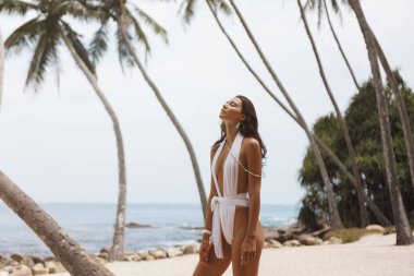 Güzel bir kadın şık beyaz bir mayo içinde sallanan palmiye ağaçları ve dingin bir okyanus zeminiyle tropikal bir plajda güven içinde poz veriyor. Seyahat, moda ya da yaşam tarzı temaları için mükemmel.
