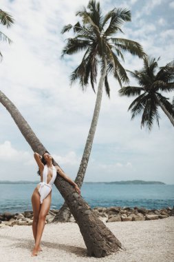 Şık beyaz bikinili kendine güvenen bir kadın, sakin bir okyanus zeminli bir palmiye ağacına yaslanıyor. Moda ve seyahat temaları için lüks, yaz tarzı ve tropikal tatilleri vurguluyor. 