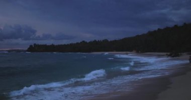 Alacakaranlıkta sakin bir tropikal plaj, hafif dalgalar, karamsar bulutlu bir gökyüzü ve uzak bir ufuk. Barışçıl, atmosferik ve seyahatle ilgili konular için ideal.