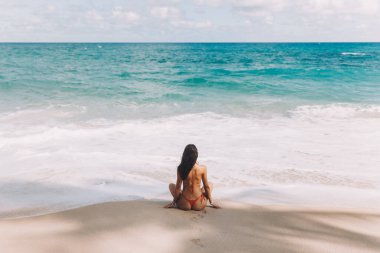 Rear view of a woman in a red bikini sitting on a sandy tropical beach, gazing at the turquoise waves. Perfect for travel, summer, and serene lifestyle-themed campaigns. clipart