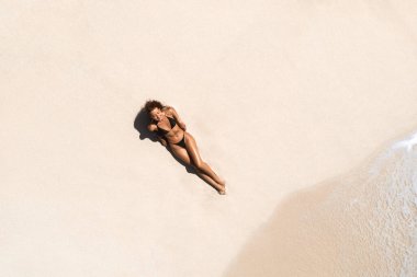 Minimalist aerial shot of a woman in a black bikini lying on pristine white sand by the shoreline of a tranquil beach in the Maldives. Perfect for travel, summer, and relaxation themes.  clipart