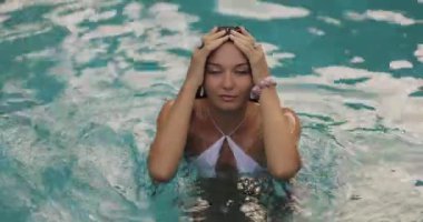 Beyaz bikinili bir kadın tropik bir havuzda dikilirken parmaklarını saçlarında gezdirir. Ağır çekimde yakalanan bu yüksek kaliteli stok videosu rahatlama, yaz ve