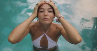 Beyaz bikinili bir kadın tropik bir havuzda duruyor, iki eliyle başını tutuyor ve kameraya bakıyor. Bu yüksek kaliteli stok görüntüleri ferahlatıcı ve huzurlu bir anı yakalar.