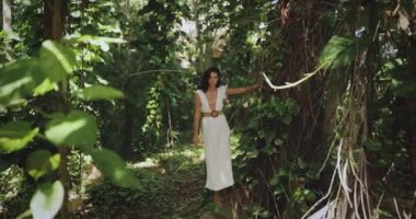 Koyu yakalı zarif beyaz elbiseli bir kadın gür tropikal bir ormanı keşfediyor, eli nazikçe yeşilliklere dokunuyor. Bu yüksek kaliteli stok videosu maceranın ve modanın özünü yakalıyor.