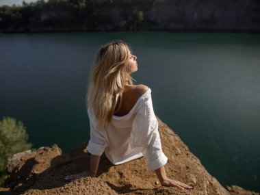 Uçurumun kenarında oturan, güneşin tadını çıkaran ve sakin göl manzarasının tadını çıkaran sarışın bir kadının huzurlu bir görüntüsü. Bu yüksek kaliteli stok fotoğrafı rahatlama, doğal güzellik ve...