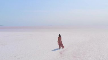 Havadan manzaralı pembe bir tuz gölünde yürüyen şık bir kadının elbisesi rüzgarda sallanıyordu. Seyahat, moda veya minimalist doğa görselleri için ideal. 