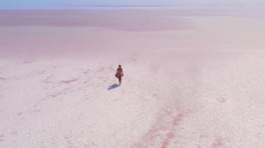 Parlak bir gökyüzünün altında, engin pembe bir tuz gölünde tek başına yürüyen bir kadının büyüleyici bir görüntüsü. Huzurlu ve minimalist bir manzara yaratıyor. Seyahat ve doğa temaları için ideal. 