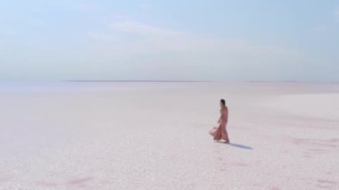 Uçsuz bucaksız pembe bir tuz gölünde berrak mavi bir gökyüzünün altında zarif bir şekilde yürüyen, akışkan elbiseli bir kadının büyüleyici bir görüntüsü. Seyahat, doğa ve moda temaları için mükemmel.. 
