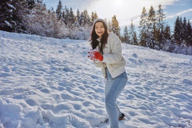 Sıcak kış kıyafetleri içinde neşeli genç bir kadın arka planda güneş ışığı ve ormanla karlı bir tarlada bir kartopu tutuyor. Yaşam tarzı, eğlence, tatil veya mevsimlik temalar için mükemmel. 