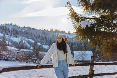 Kışın giyinmiş genç bir kadın, karlı bir kırsal alanda ahşap bir çite yaslanmış, sakin kış manzarasının tadını çıkarıyor..